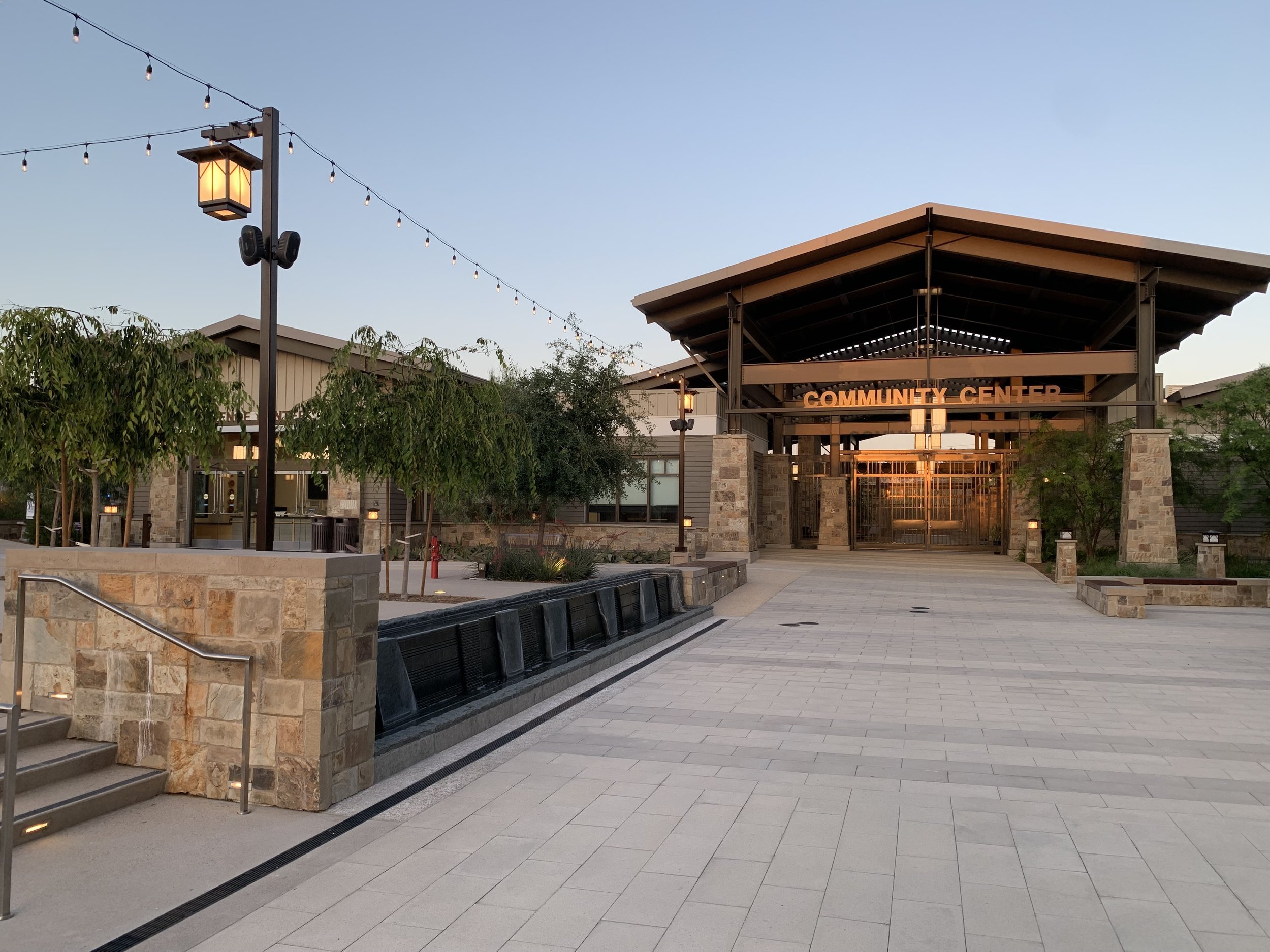 Entrance of a modern civic center with stone pillars, large open pavilion, string lights, and surrounding greenery.