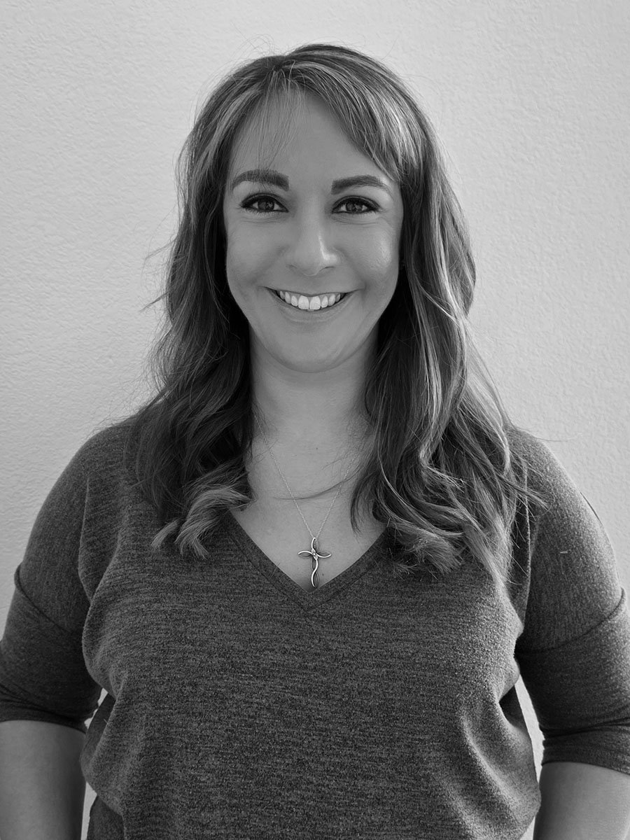 Smiling woman with long hair wearing a gray V-neck top and a cross necklace, standing against a white background.